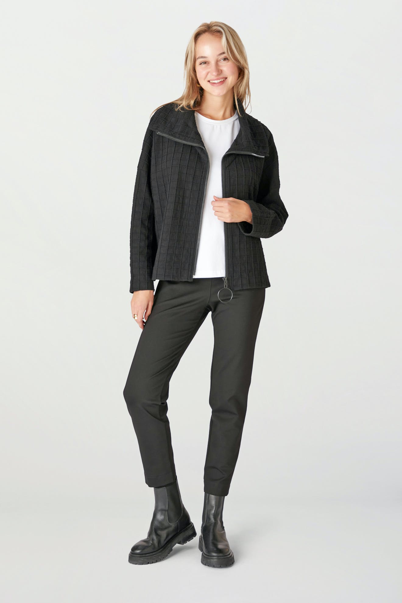 A person wearing a black Window Pane Wool Jacket featuring an oversized folded collar, paired with a white shirt, black pants, and boots stands against a light background. They are smiling and posing casually.