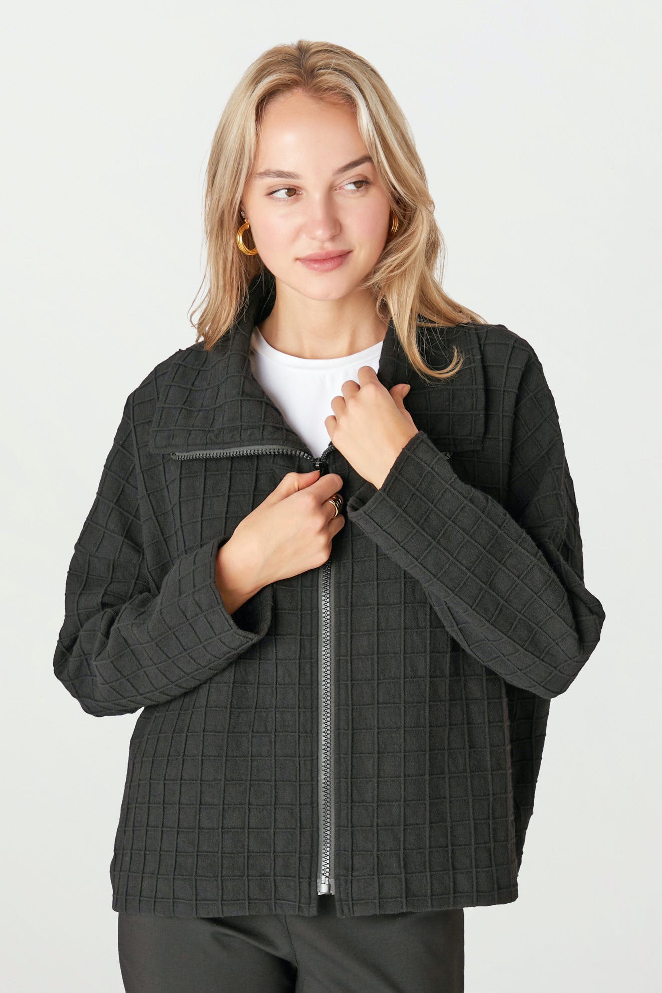 A person with blonde hair is wearing a Window Pane Wool Jacket in black over a white top and black pants. They are holding the open zipper of the structured jacket and looking slightly to the side.