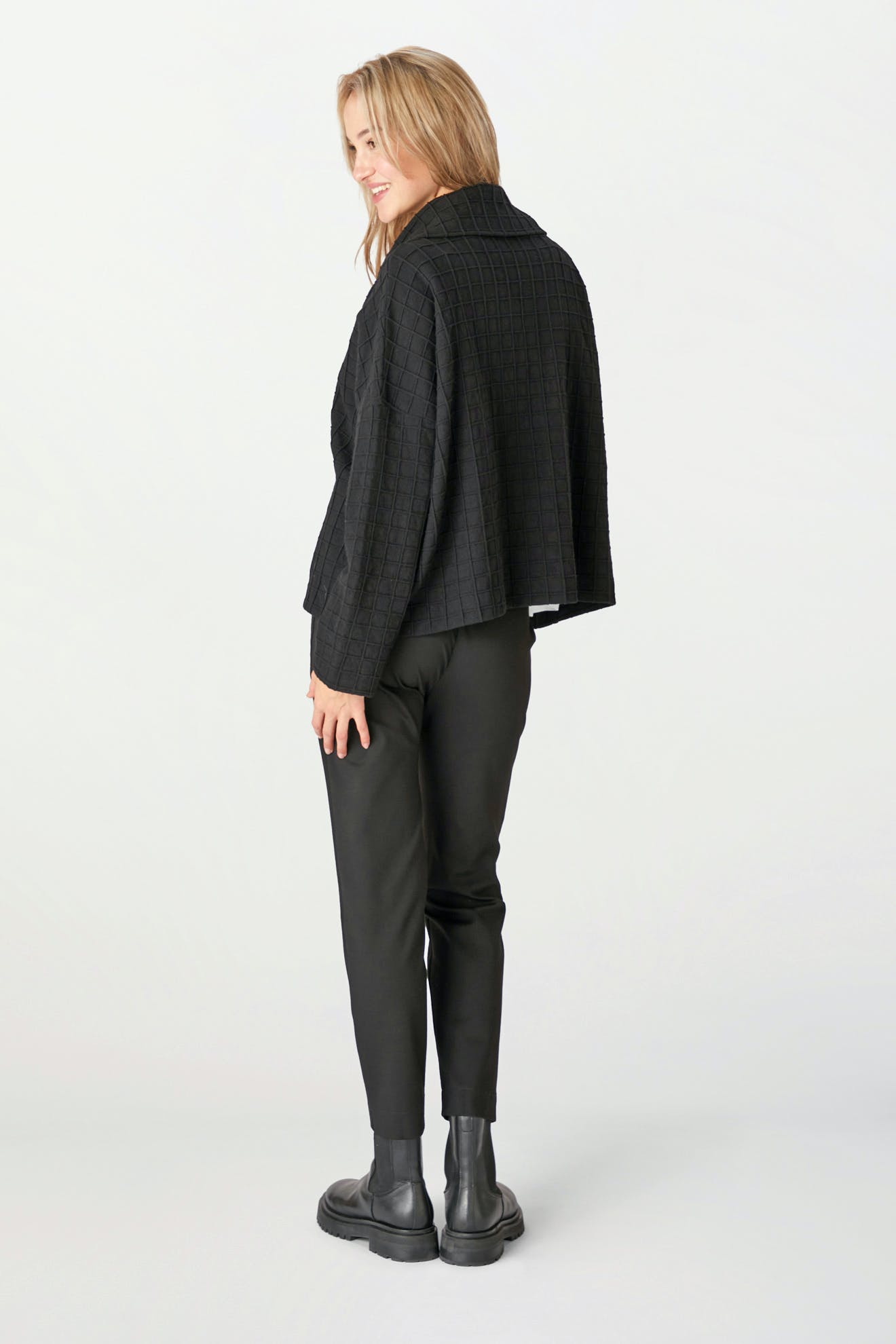 A person wearing the Window Pane Wool Jacket, made from a black cotton wool blend with an oversized folded collar, pairs it with black pants and boots while standing slightly sideways against a light background.