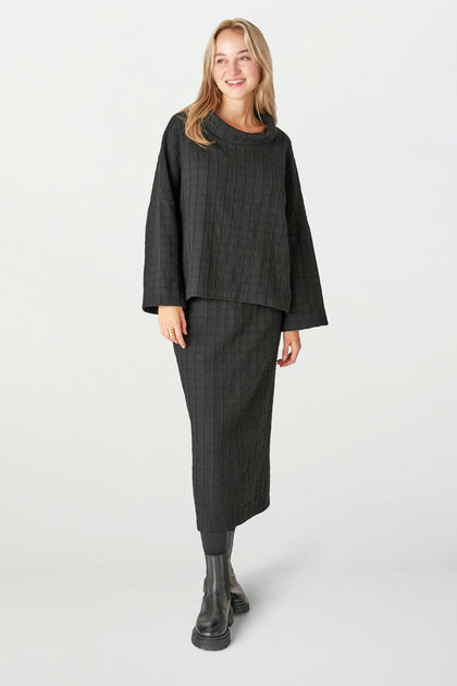 A woman faces forward, wearing the Window Pane Wool Top—a black wool piece with a relaxed turtleneck and boxy cut—paired with a matching long skirt and black boots, set against a plain light background.