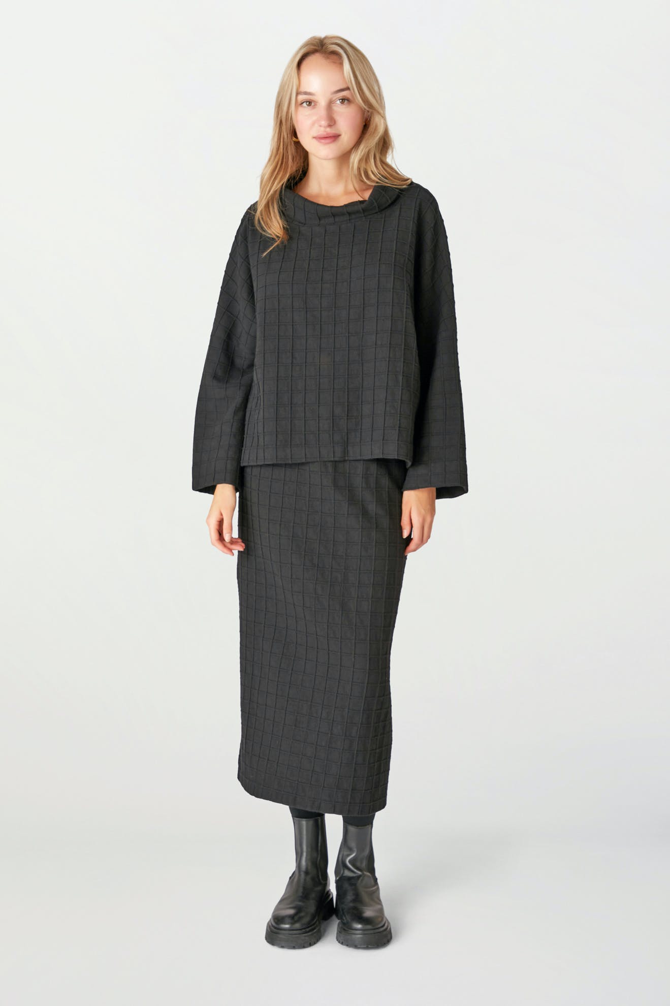 A woman stands against a plain background wearing a relaxed black Window Pane Wool Top, a matching boxy cut black quilted skirt, and black boots.