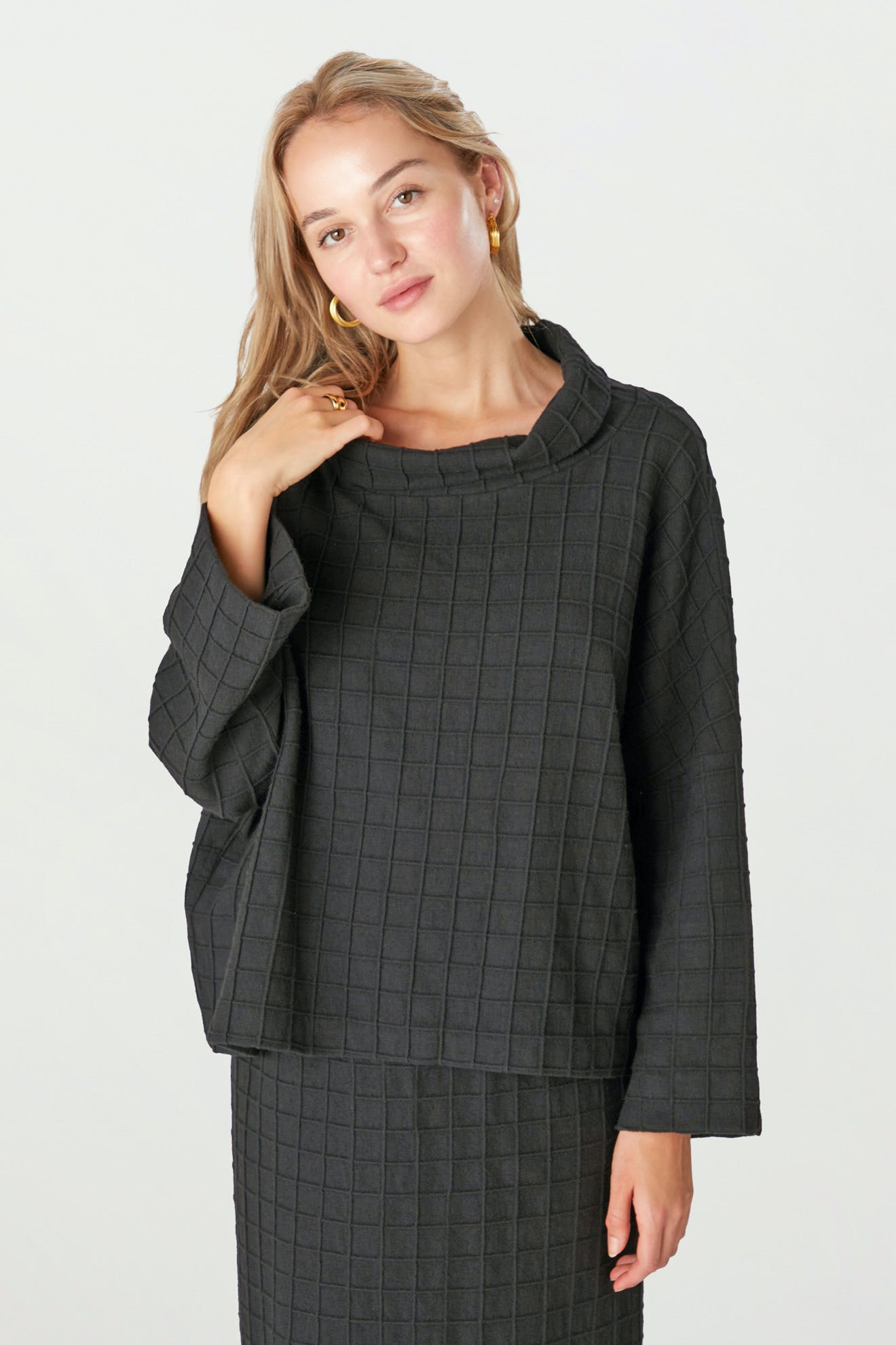 A woman wears the Window Pane Wool Top, a loose-fit, boxy black long-sleeve top, paired with a matching skirt against a plain light background.