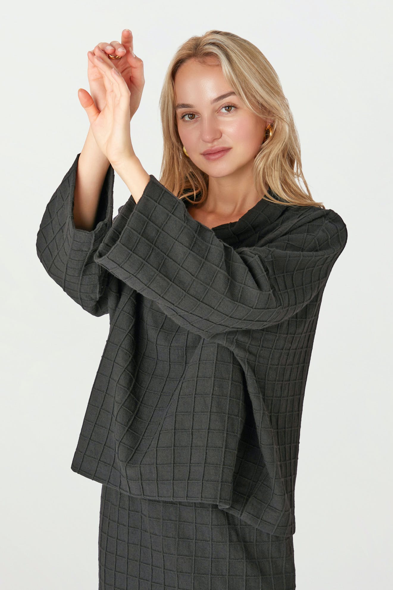 A woman in a Window Pane Wool Top featuring a dark grid pattern and boxy cut stands with arms raised and crossed, gazing at the camera against a plain background.