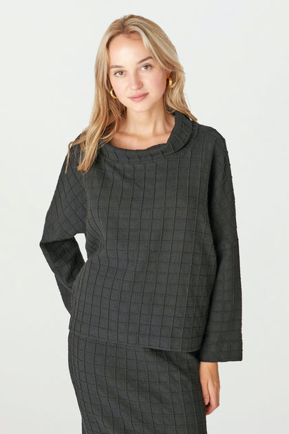 A woman wears the Window Pane Wool Top—a dark, textured wool piece with a relaxed turtleneck and boxy cut—paired with a matching skirt, posed against a plain light background.