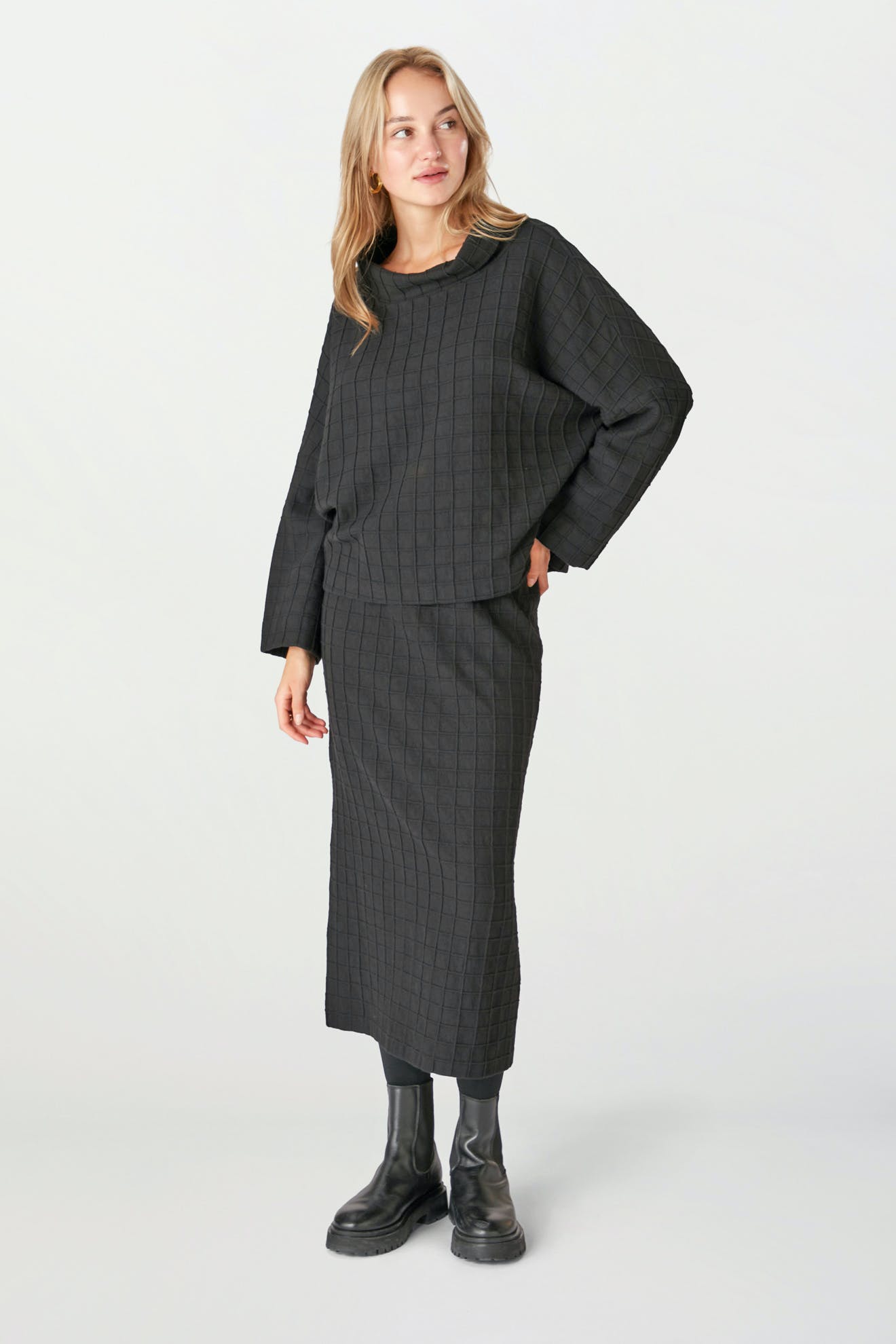 A person wearing the Window Pane Wool Top, a black textured wool top with long sleeves and a relaxed turtleneck, paired with a matching skirt and black boots, stands against a plain light background.