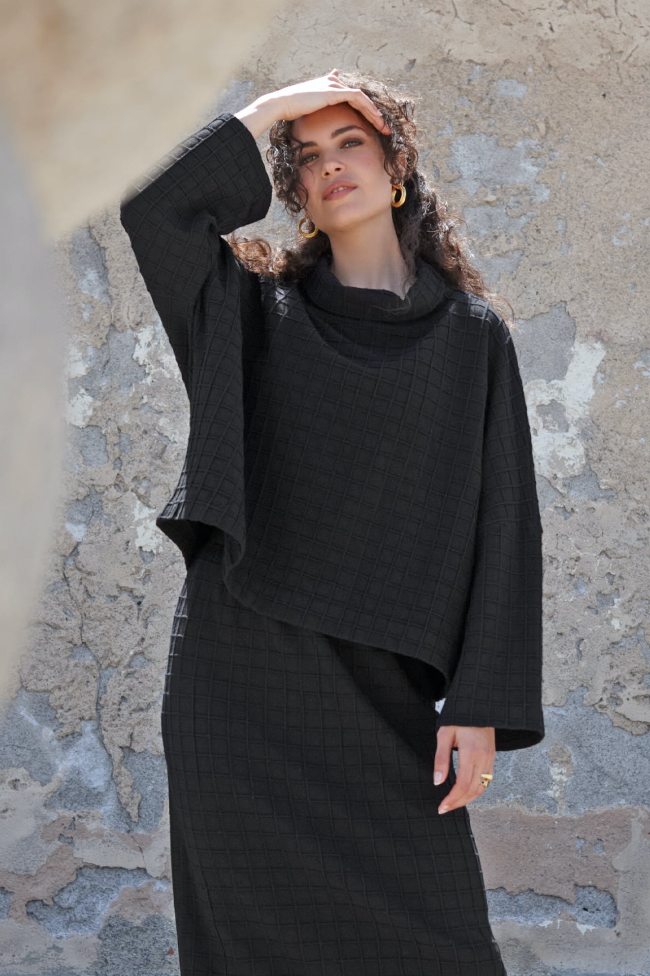 A woman in a boxy-cut Window Pane Wool Top stands by a weathered stone wall, shielding her eyes from the sun with one hand.
