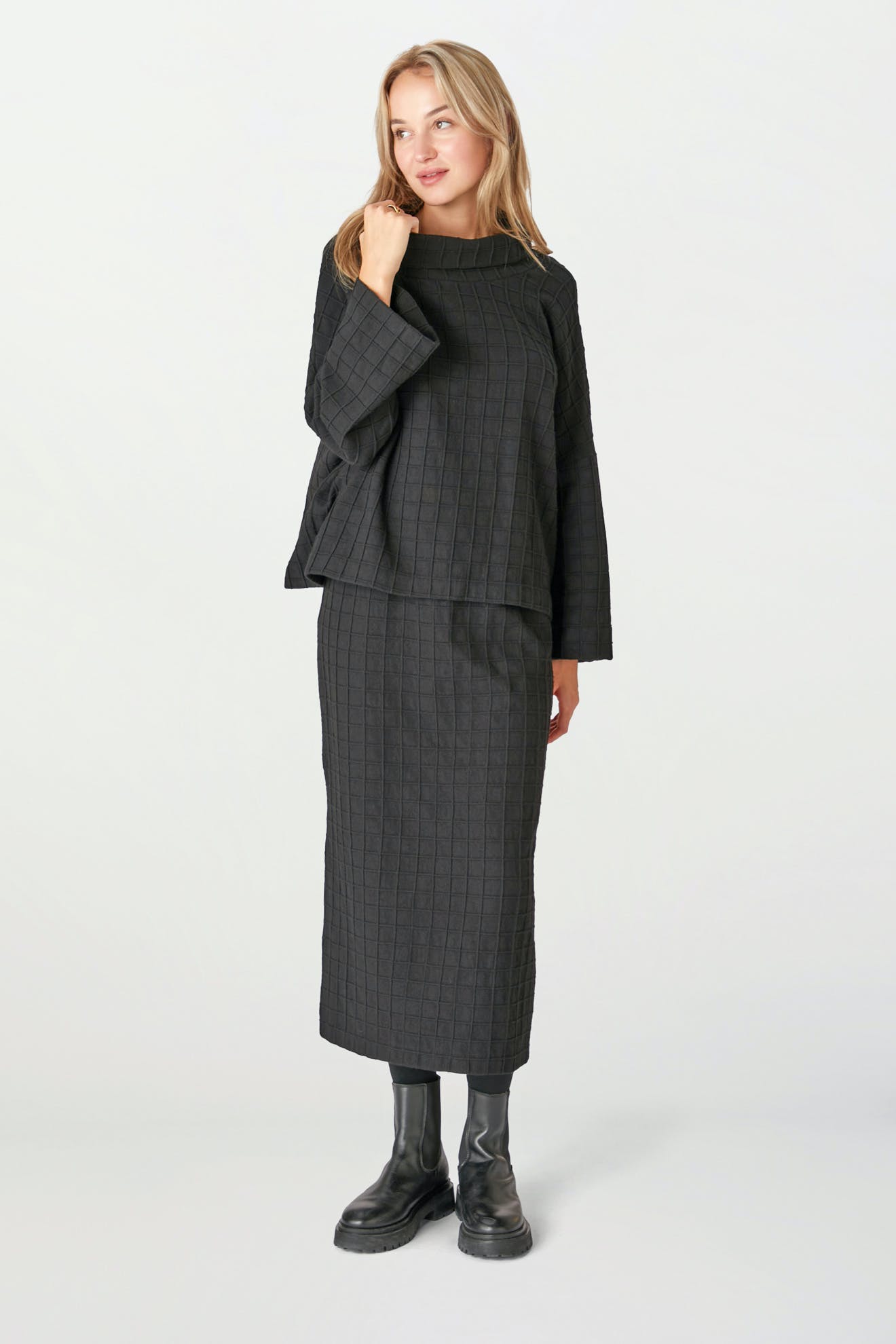 A person stands against a plain light background wearing the Window Pane Wool Top— a black, checkered, boxy-cut long-sleeve top—paired with a matching ankle-length skirt and black boots.