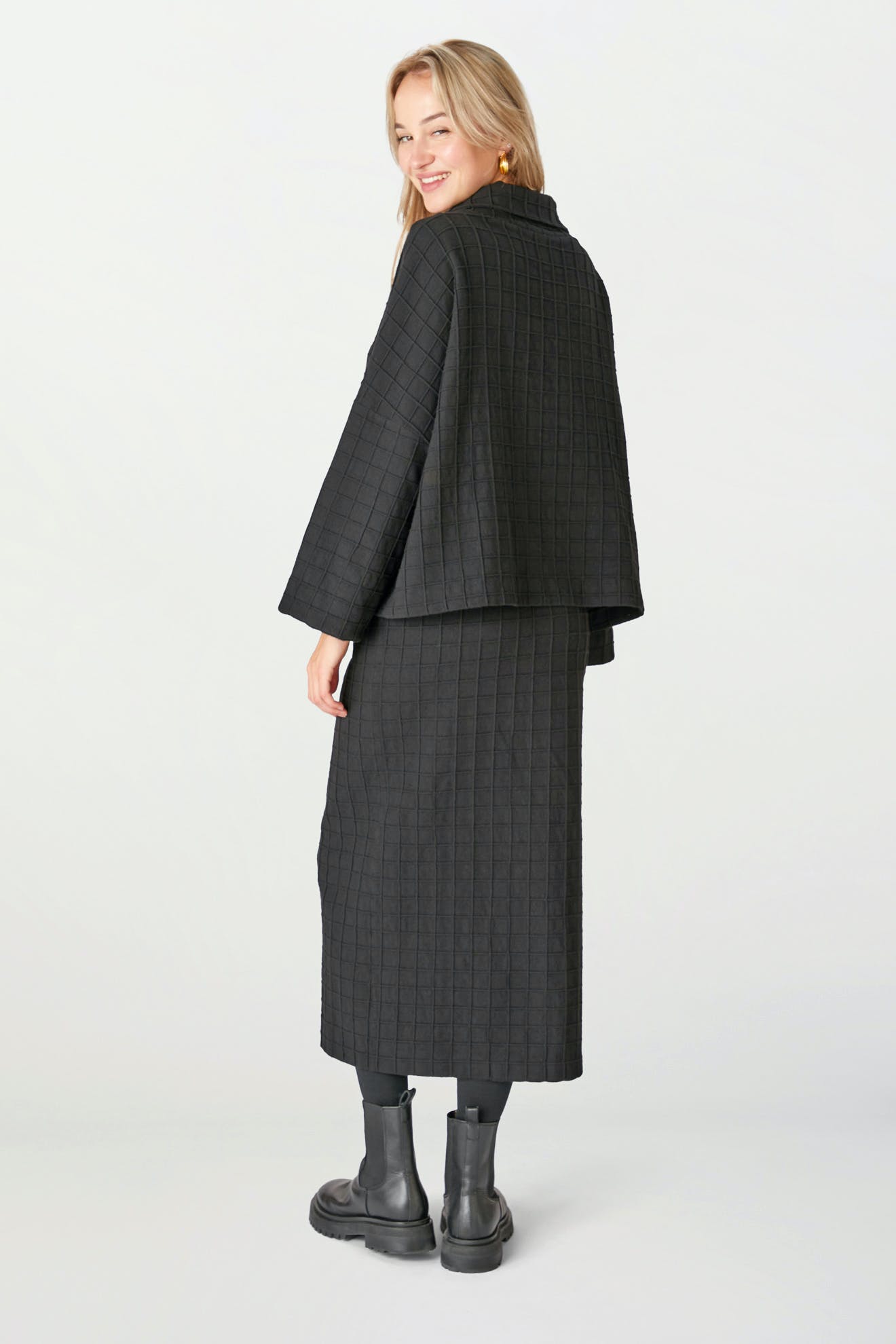A person stands facing slightly away, wearing the Window Pane Wool Top with a boxy cut, paired with a matching long skirt, black boots, and gold hoop earrings.