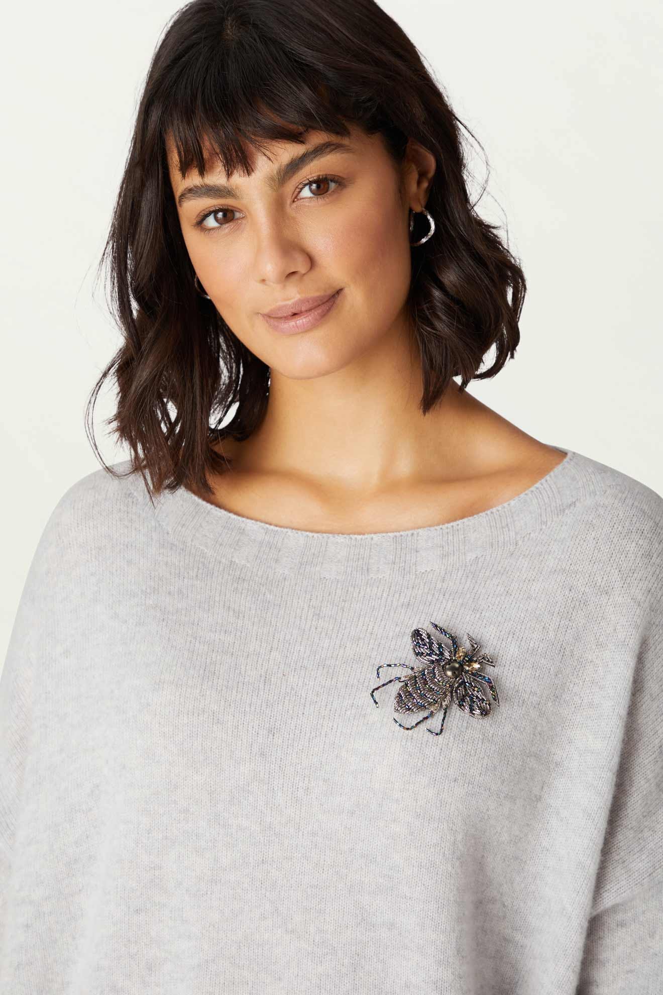 Woman wearing a light grey sweater adorned with the Embroidered Bee Brooch on the left side of her chest, gazing at the camera.