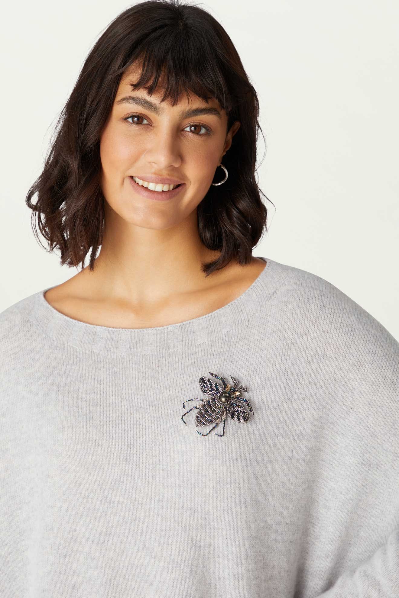 A woman smiles at the camera, wearing a light gray sweater adorned with the Embroidered Bee Brooch pinned on the left side.
