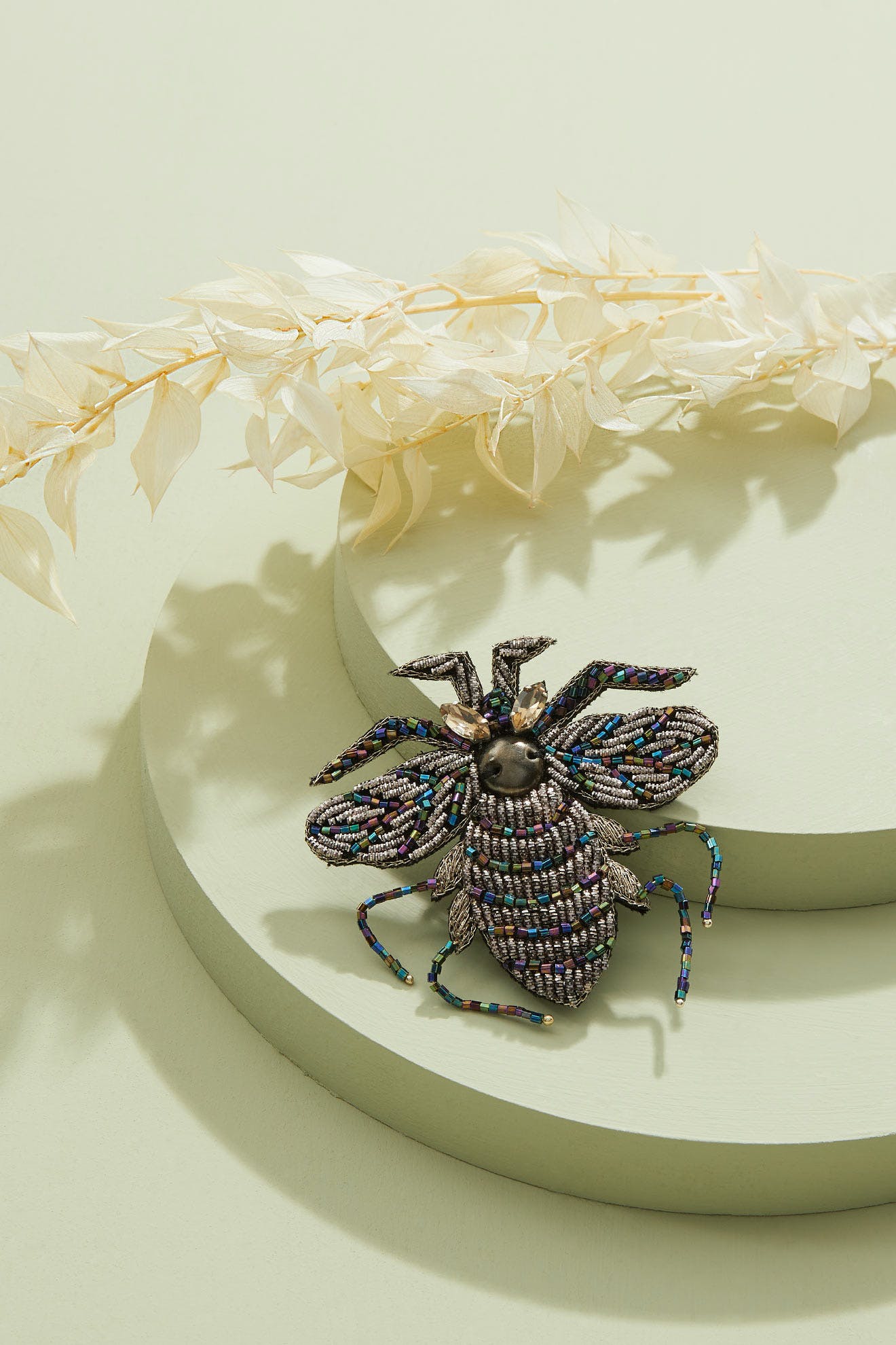 Embroidered Bee Brooch with intricate details rests on light green round platforms beside cream-colored dried leaves.