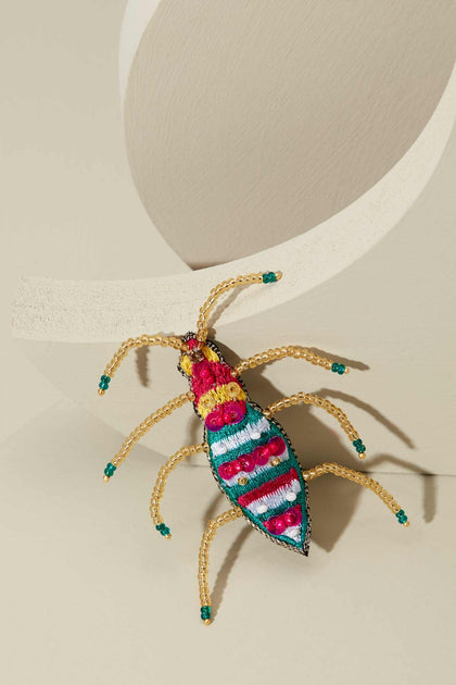The Sequin Detail Brooch, a beetle-shaped accessory adorned with vibrant seed beads and intricate embroidery, is elegantly displayed on a beige curved surface.