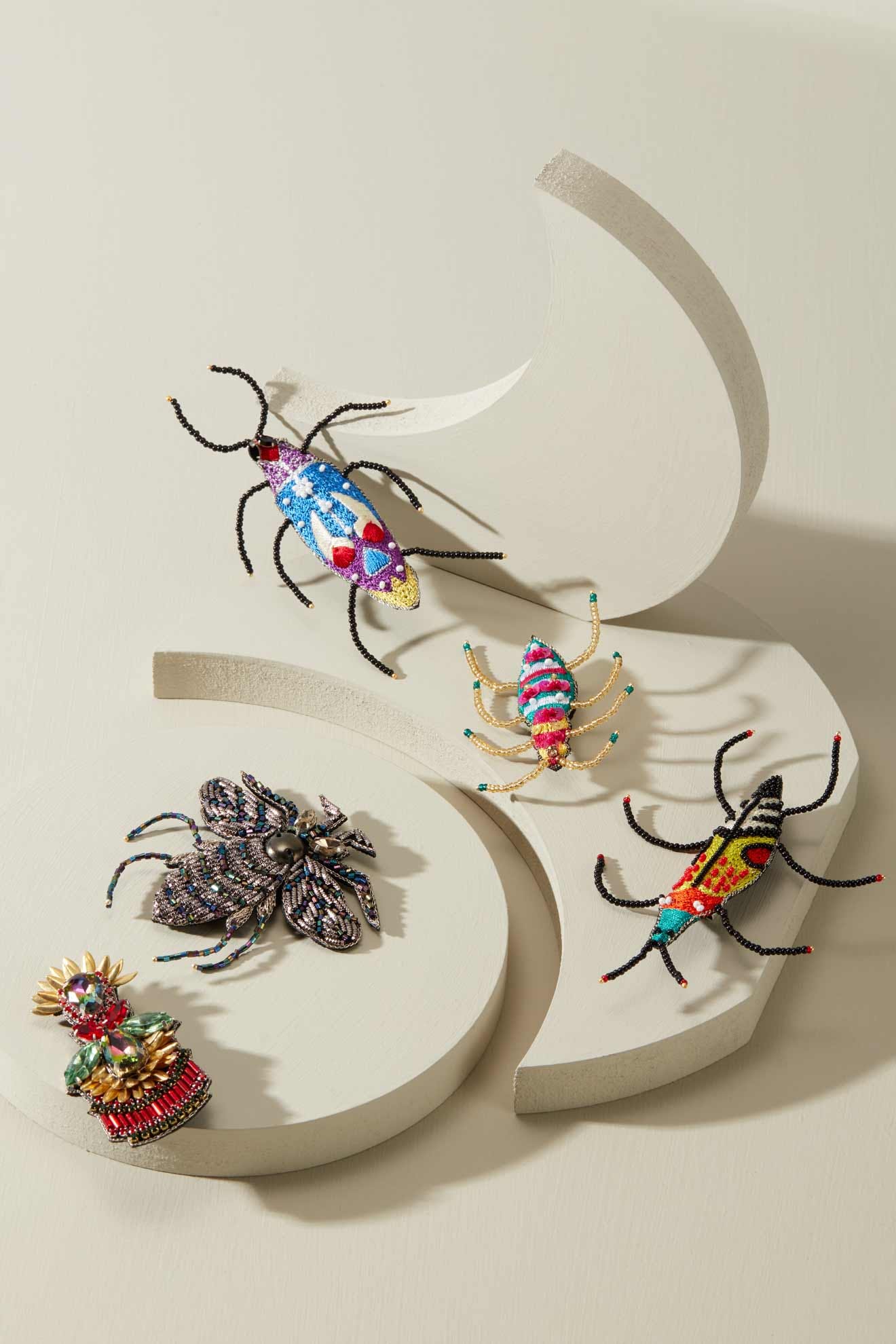 A display of Sequin Detail Brooches featuring colorful, beaded insects such as beetles, spiders, and a bee, expertly crafted with vibrant seed beads and intricate embroidery, all elegantly arranged on a white geometric stand.