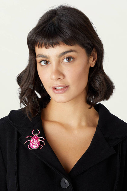 A woman with short dark hair wears a black jacket decorated with a Pink Detail Beetle Brooch on the lapel.