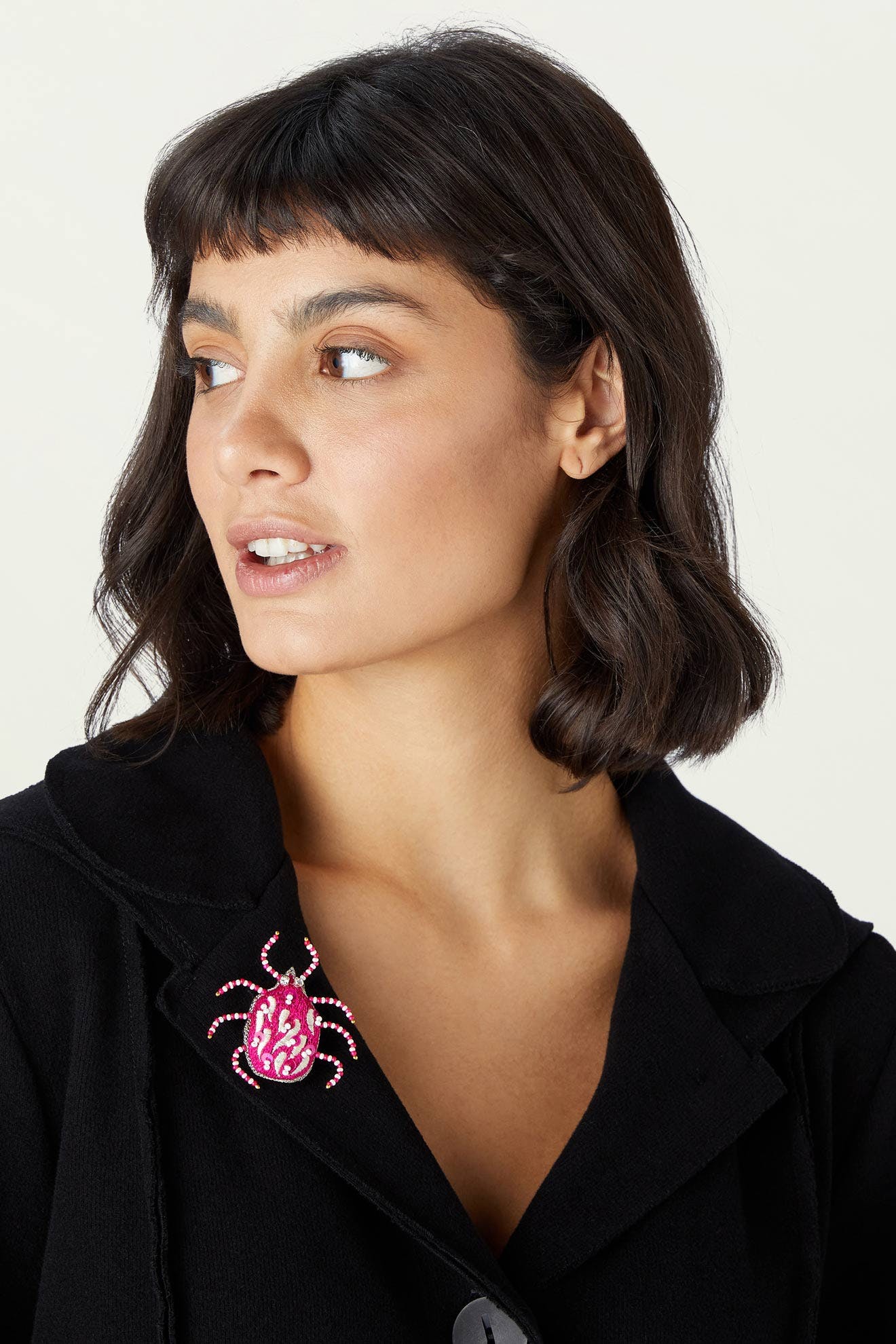 A woman with short dark hair wears a black jacket adorned with the Pink Detail Beetle Brooch on the collar, looking to the side against a plain background.