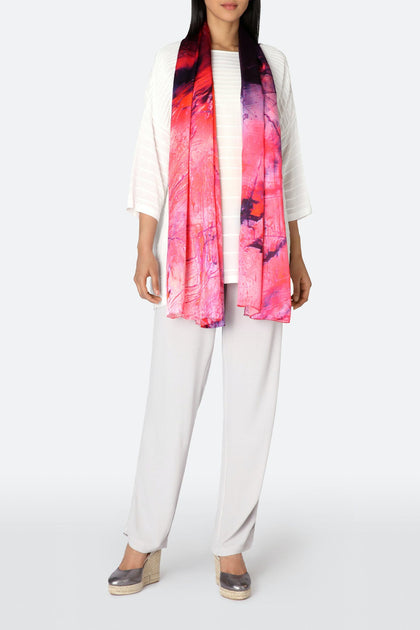 A person wearing a white outfit and silver shoes stands with the vibrant Heavenly Calcite Scarf, draped around their neck in shades of pink and purple silk, showcasing impeccable fashion sense.