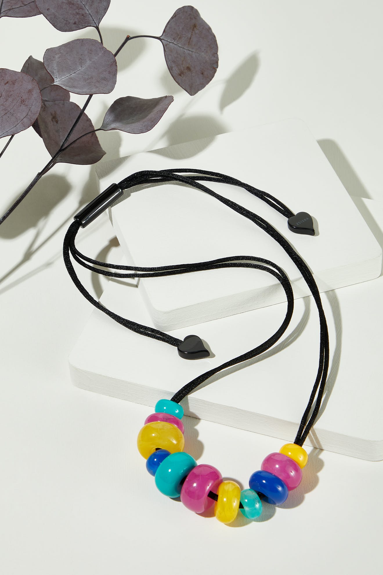 The Kaleidoscopic Necklace, featuring large resin beads in yellow, blue, pink, and purple on an adjustable black cord, is displayed on a white surface beside dried leaves—a vibrant choice for handmade jewelry lovers.