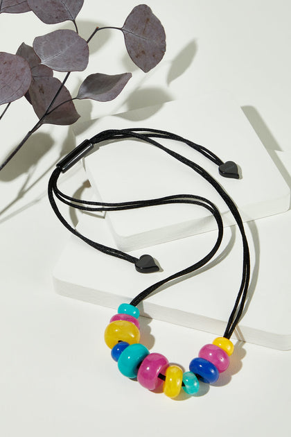 The Kaleidoscopic Necklace, featuring large resin beads in yellow, blue, pink, and purple on an adjustable black cord, is displayed on a white surface beside dried leaves—a vibrant choice for handmade jewelry lovers.