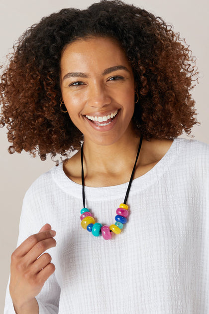 A woman with curly hair, smiling at the camera, wears a white textured top and the Kaleidoscopic Necklace.