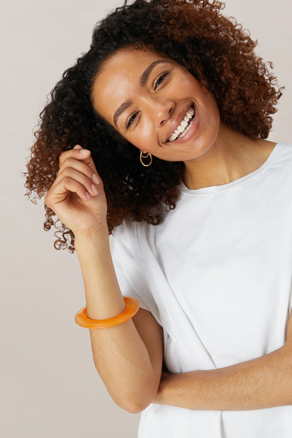 A woman with curly hair smiles against a light background, wearing a white shirt, gold hoop earring, and the Marbelled Bracelet on her wrist.