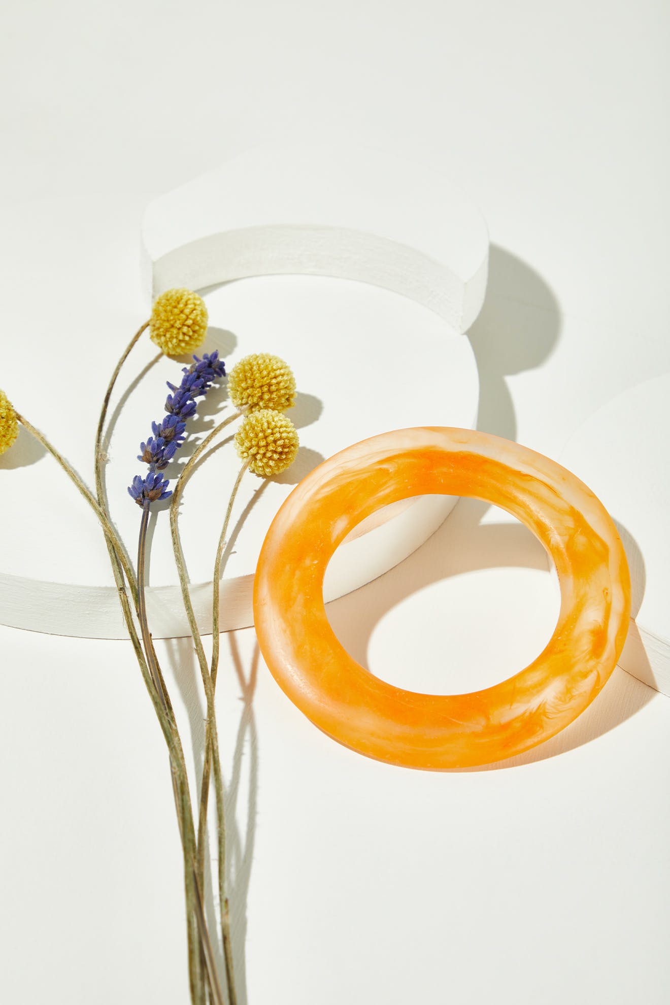 The Marbelled Bracelet, featuring orange marbled resin, is displayed beside dried yellow and purple flowers on a white surface with round platforms.