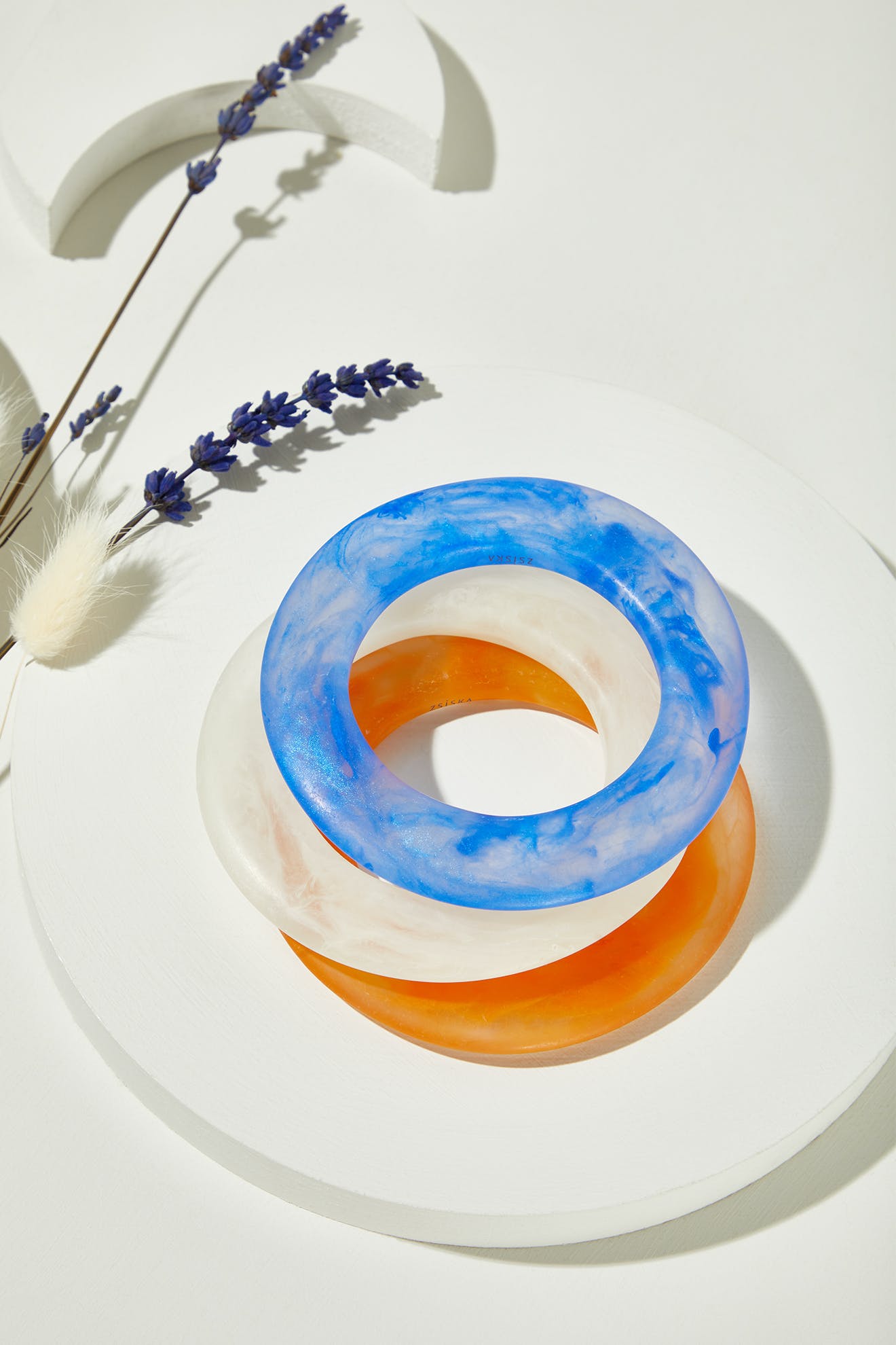 The Marbelled Bracelet, a set of three resin bangles in blue, white, and orange, is displayed stacked on a round white platform beside sprigs of dried lavender.