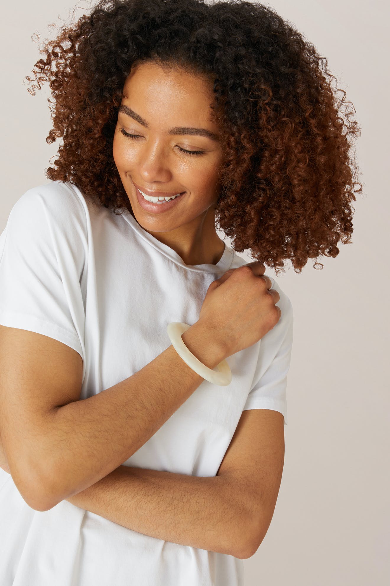 A woman with curly hair in a white t-shirt smiles while looking down, one arm crossed to show off her Marbelled Bracelet.