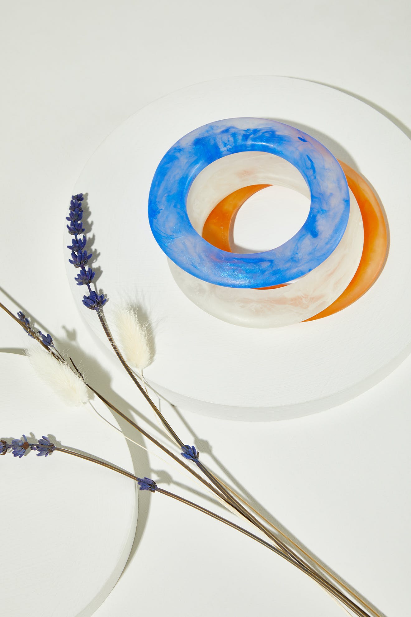 The Marbelled Bracelet, available in blue, orange, and clear resin, features a translucent marbled design. Displayed on white platforms with dried lavender and grasses, these bangles add unique charm to any handmade jewelry collection.