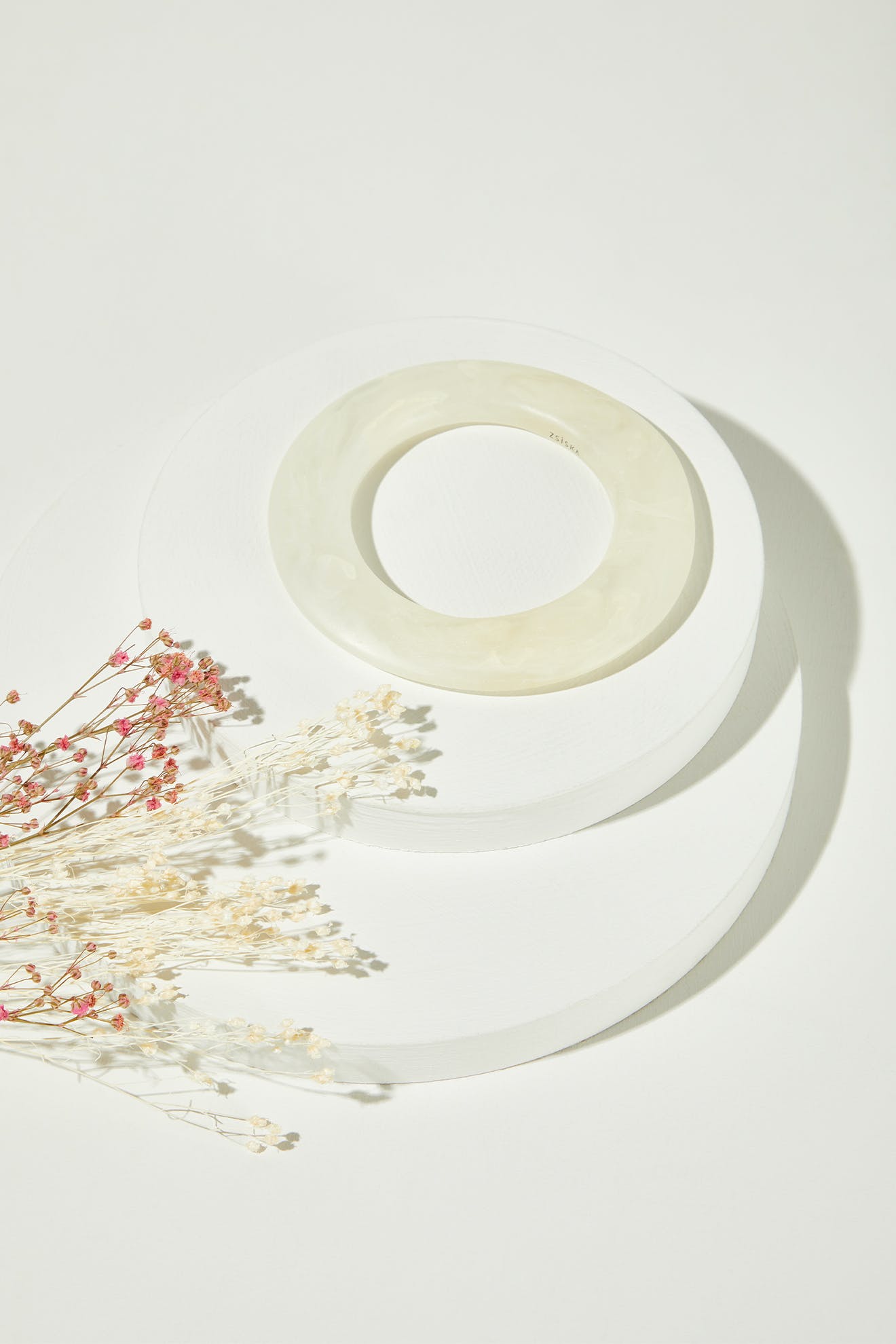 The Marbelled Bracelet rests on stacked white circular platforms beside dried white and pink flowers.